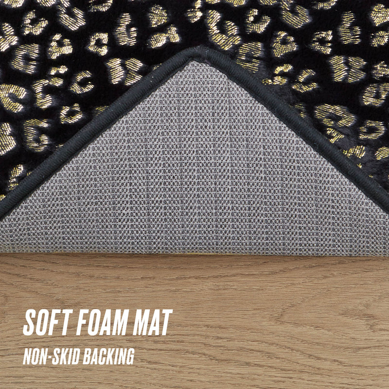 A corner of the Metallic Leopard Plush Rug, 3' Heart with leopard print is flipped to reveal its gray non-skid backing. The soft foam mat sits on a wooden floor. Text: Soft Foam Mat, Non-Skid Backing.