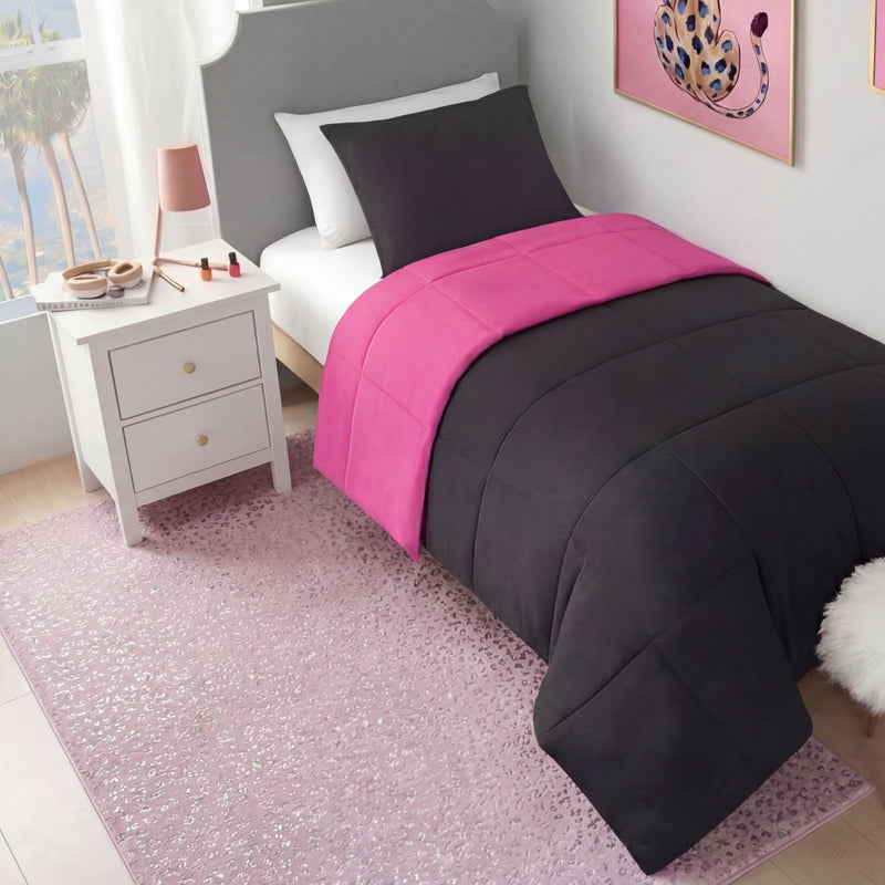 A neatly made twin bed with a black and bright pink comforter sits beside a white nightstand holding a lamp and makeup. The room features pink decor, a Metallic Leopard Plush Rug (4' x 6'), and a window with palm trees outside.