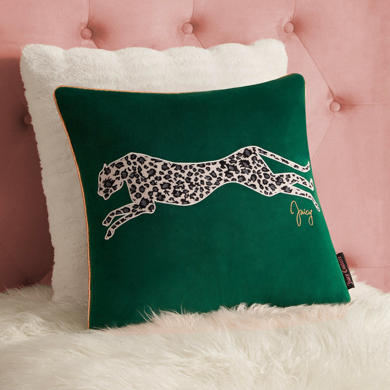 The Velvet Cheetah Square Pillow features a green velvet cover with a cheetah applique and “Juicy” in gold script, placed before a white fluffy pillow on a pink tufted background over white faux fur.