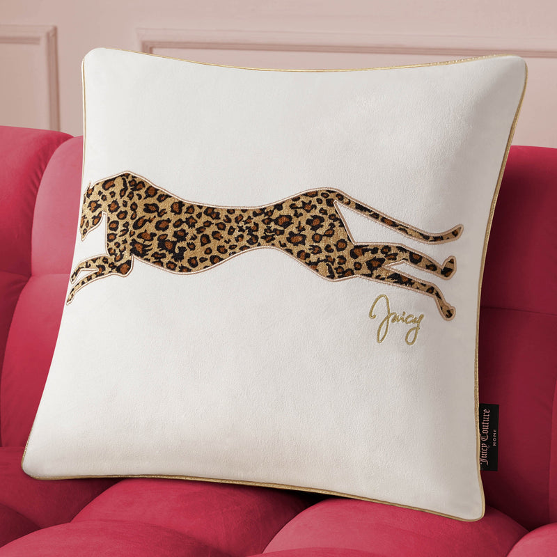 The Velvet Cheetah Pillow, featuring gold piping, an embroidered cheetah, and "Juicy" in gold script, adds a stylish Juicy Couture accent to any space when placed on a red tufted sofa.