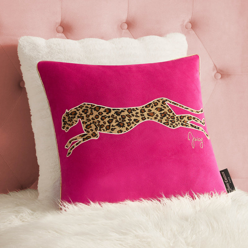 A bright pink Velvet Cheetah Square Pillow with a leopard-print cheetah applique and "Juicy" embroidery rests on white faux fur, set against a white pillow and a pink upholstered wall.