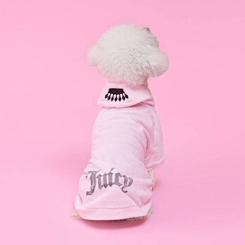 A small white dog sits facing away against a solid pink background, dressed in the Bling Velour Pet Hoodie—a light pink Juicy Couture hoodie featuring sparkling rhinestone “Juicy” lettering and an eyelash design on the hood—showcasing adorable designer pet fashion.