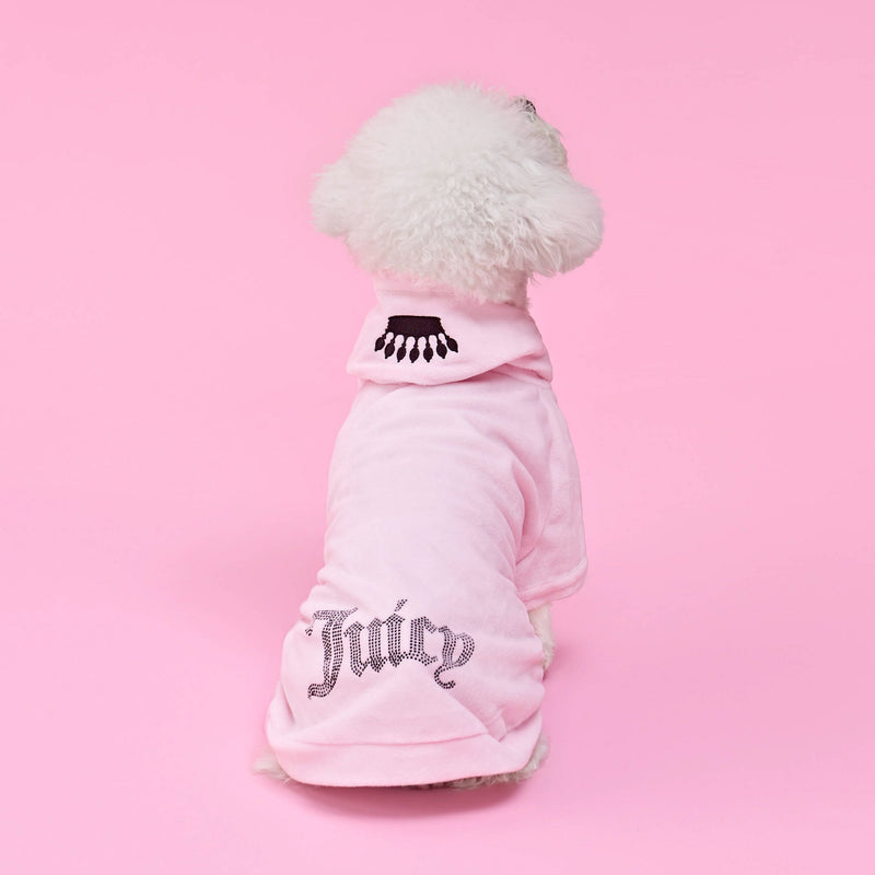 A small white dog sits facing away against a solid pink background, dressed in the Bling Velour Pet Hoodie—a light pink Juicy Couture hoodie featuring sparkling rhinestone “Juicy” lettering and an eyelash design on the hood—showcasing adorable designer pet fashion.