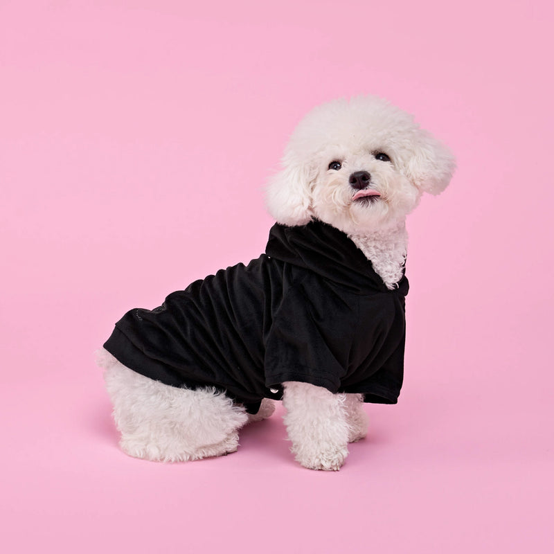 A fluffy white dog wearing a Bling Velour Pet Hoodie sits on a pink background, looking slightly to the side with its tongue out.