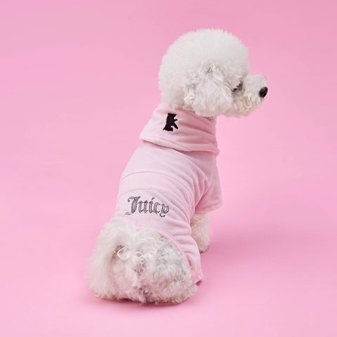A fluffy white dog faces away from the camera, sitting on a pink background and modeling the Bling Velour Pet Tracksuit in light pink, which features “Juicy” written on the back.