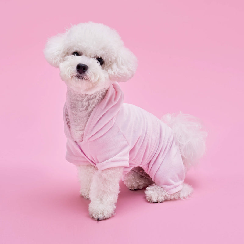 A fluffy white poodle wearing the Bling Velour Pet Tracksuit in light pink sits against a solid pink background, looking toward the camera and showcasing the latest in pet fashion.