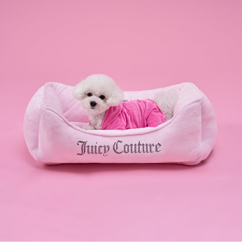 A small white dog wearing a pink outfit sits in a plush black Crown Bling Pet Bed with Juicy Couture written on the front, all against a pink background.