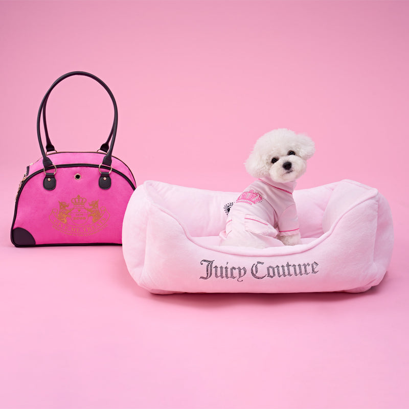 A small white dog wearing a pink outfit sits in a Crown Bling Black Pet Bed with sparkling accents, next to a matching Juicy Couture handbag, all set against a pink background.