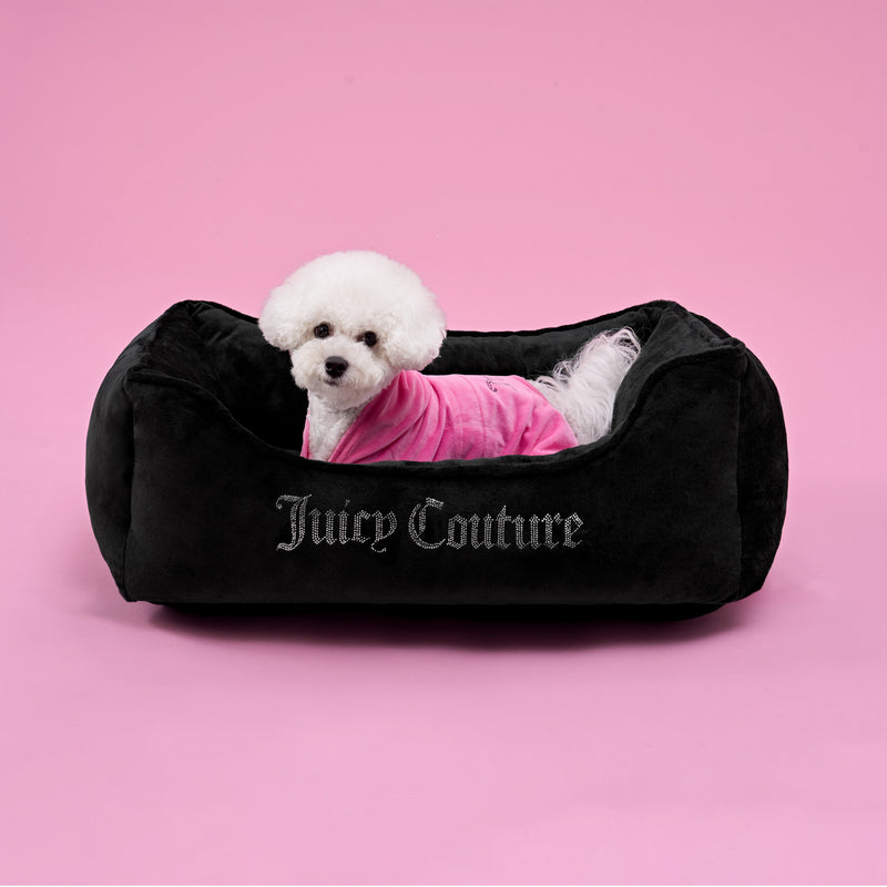 A small white dog in a pink outfit sits on a luxurious Crown Bling Black Pet Bed against a solid pink background—the perfect spot for small to medium pets to relax in style.