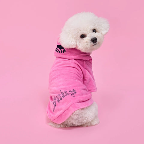 A fluffy white dog wears the Bling Velour Pet Hoodie—a pink hooded robe with a black eyelash design and glittery text—while sitting on a pink background and looking over its shoulder, the perfect touch of designer pet apparel.