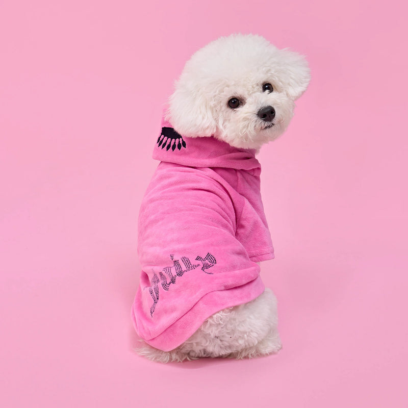 A fluffy white dog wears the Bling Velour Pet Hoodie—a pink hooded robe with a black eyelash design and glittery text—while sitting on a pink background and looking over its shoulder, the perfect touch of designer pet apparel.