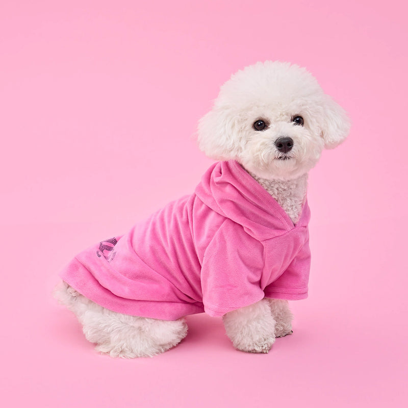 A fluffy white dog wearing the Bling Velour Pet Hoodie sits on a pink background, looking at the camera. The chic pink hooded robe from Juicy Couture adds a touch of designer pet apparel to its adorable look.