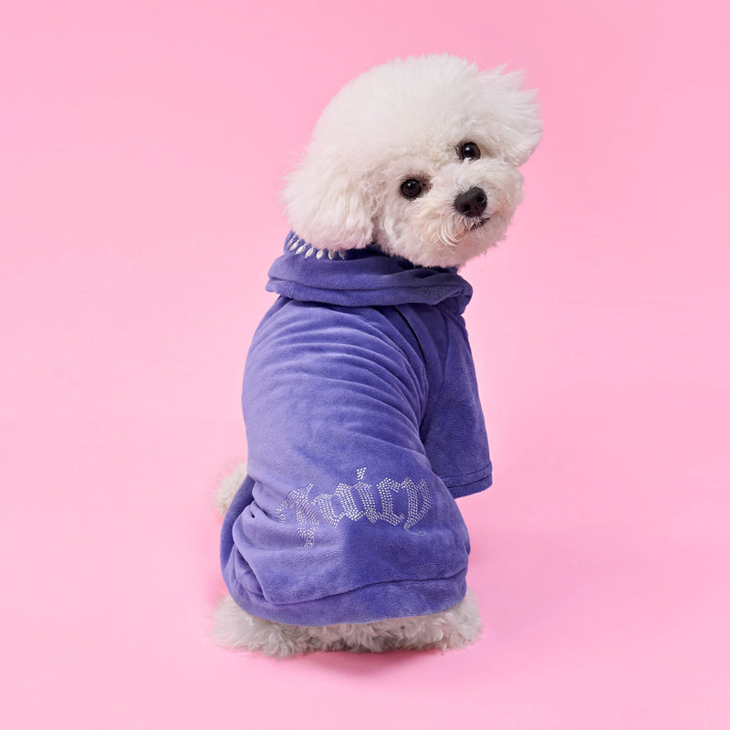 A fluffy white dog sits on a pink background, wearing a soft purple Bling Velour Pet Hoodie with silver lettering on the sleeve, looking over its shoulder toward the camera.