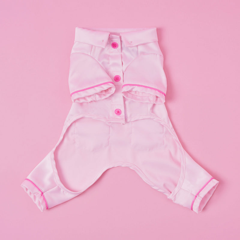 The Juicy Couture Pet Silky Satin Pajamas feature a pink silky satin fabric with short sleeves and legs, button-down front, and pink trim, displayed flat against a matching pink background to highlight their shape.