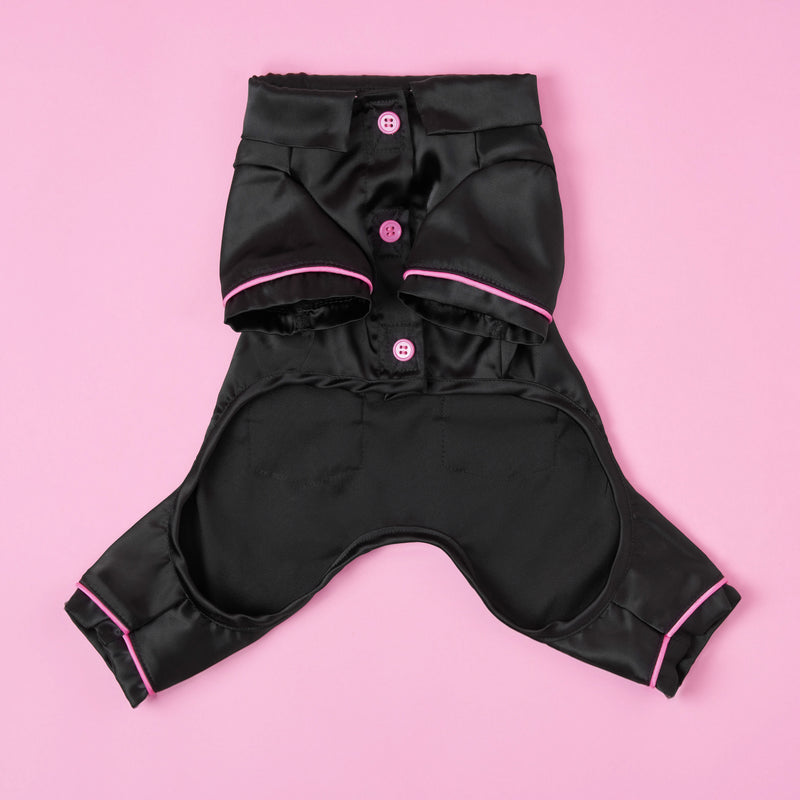 Juicy Couture Pet Silky Satin Pajamas in black with pink trim and buttons, displayed flat against a pink background. Designed for small dogs or cats, these pajamas feature openings for your pet’s legs and tail, offering a luxurious addition to your furry friend’s wardrobe.