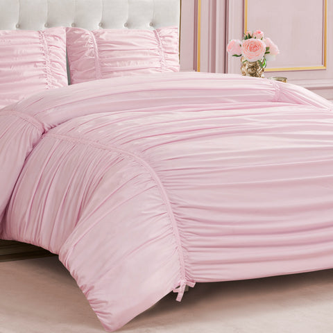 A neatly made bed features the Ribbon Ruched Comforter Set in light pink with matching pillows. In the background, there’s a pink wall with gold trim and a side table displaying a vase of pink roses.