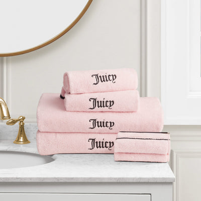 A stack of Neon Border Bath Towel Set towels in light pink with "Juicy" embroidered in black, displayed on a white marble countertop near a gold faucet and round mirror.
