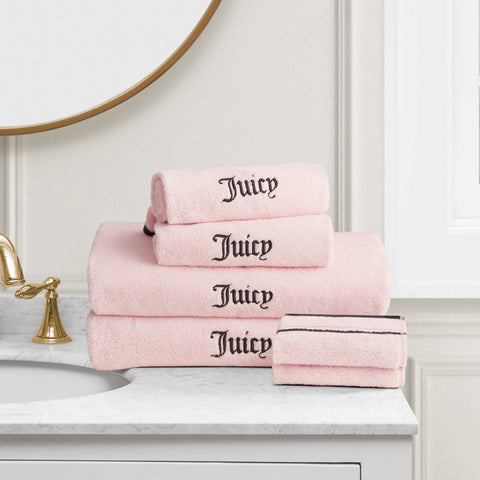A stack of Neon Border Bath Towel Set towels in light pink with "Juicy" embroidered in black, displayed on a white marble countertop near a gold faucet and round mirror.