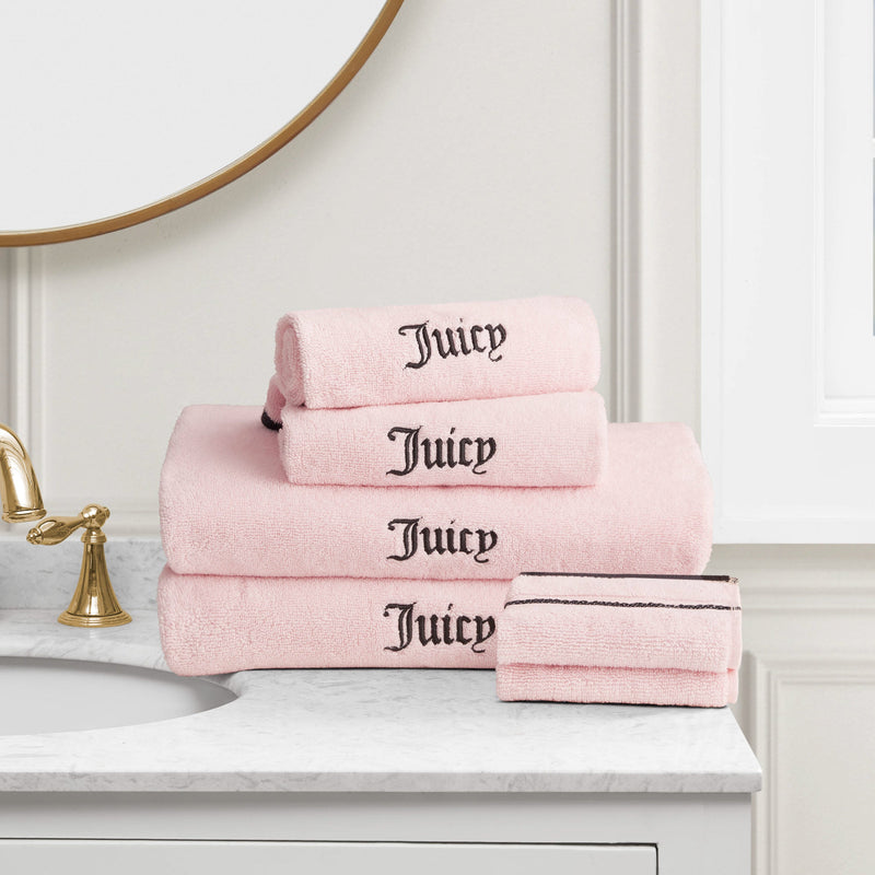 A stack of Neon Border Bath Towel Set towels in light pink with "Juicy" embroidered in black, displayed on a white marble countertop near a gold faucet and round mirror.