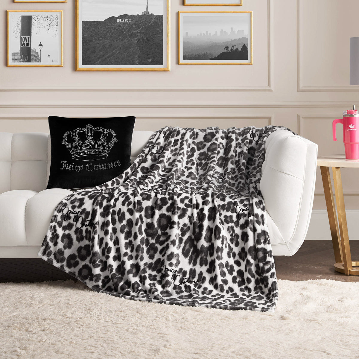 A white sofa is adorned with a black Juicy Couture pillow and an oversized Plush Throw Blanket featuring a black-and-white leopard print. On a side table sits a pink tumbler, while black-and-white photos are displayed on the wall behind.