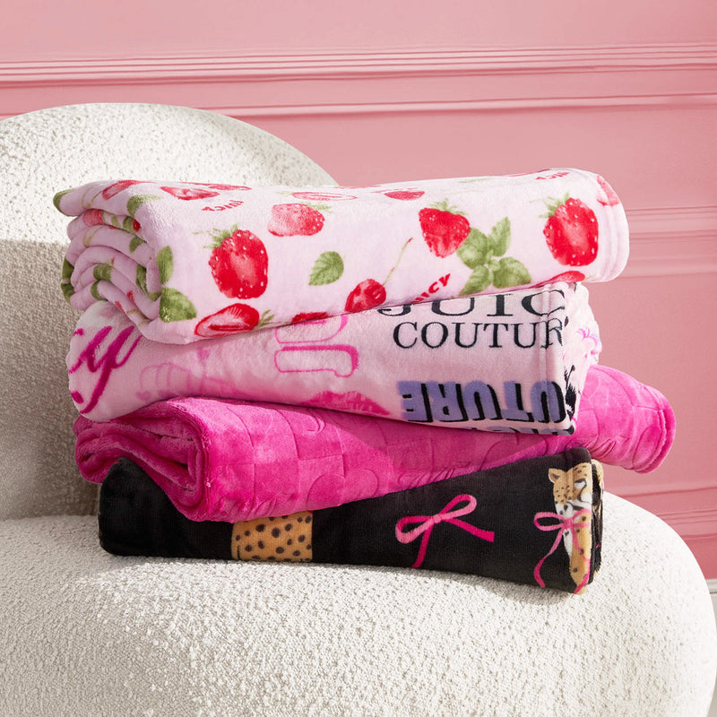 Three colorful Plush Throw Blankets are neatly folded and stacked on a textured white chair, with a pink wall in the background. The ultra-soft blankets feature strawberry, printed logo, and bow designs in pink, white, and black.