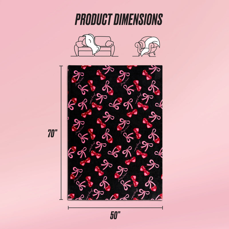 The Sweet Cherry Bows Plush Throw Blanket, in black with pink and red bow patterns, is shown on a pink background. Sized 70" x 50", this ultra-soft Juicy Couture throw is perfect for your couch or bed. Product dimensions are highlighted.