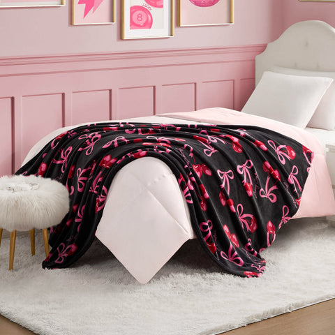 A neatly made bed with a white headboard, pink bedding, and the Sweet Cherry Bows Plush Throw Blanket showcasing pink and red bows. The room features pink walls, framed art, a fluffy white stool, and a white rug.