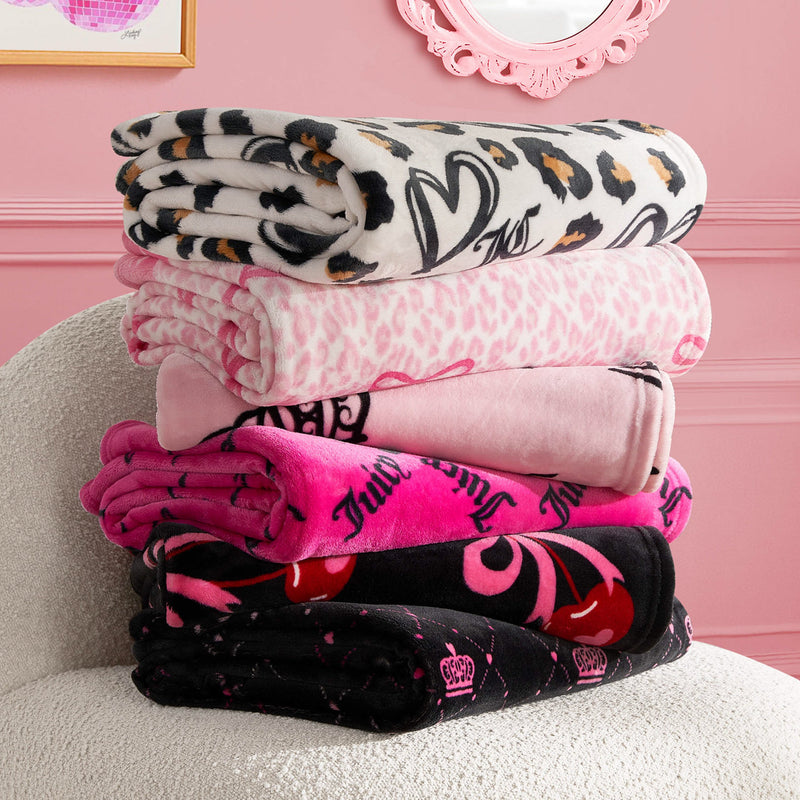 A stack of five Plush Throw Blankets in assorted pink, black, and white patterns rests on a plush white chair in a pink room with decorative frames on the wall.