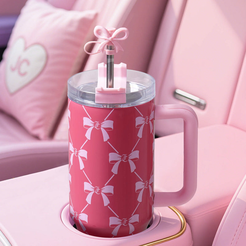 A Jetsetter Hydration Stainless Steel Tumbler, 40 oz, with a handle sits in a pastel pink car interior. The pink cup features white bows and a bow-topped straw. A heart-shaped pillow with "J C" is on the car seat in the background.