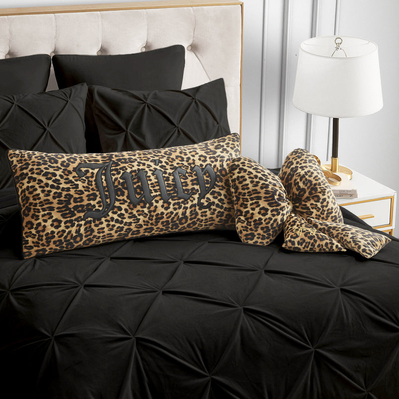 A bed with black bedding features a rectangular Juicy Couture pillow in leopard print, complemented by two Gothic Cheetah Decorative Pillows in matching cheetah print bow shapes. A white lamp sits on a nightstand next to the bed.