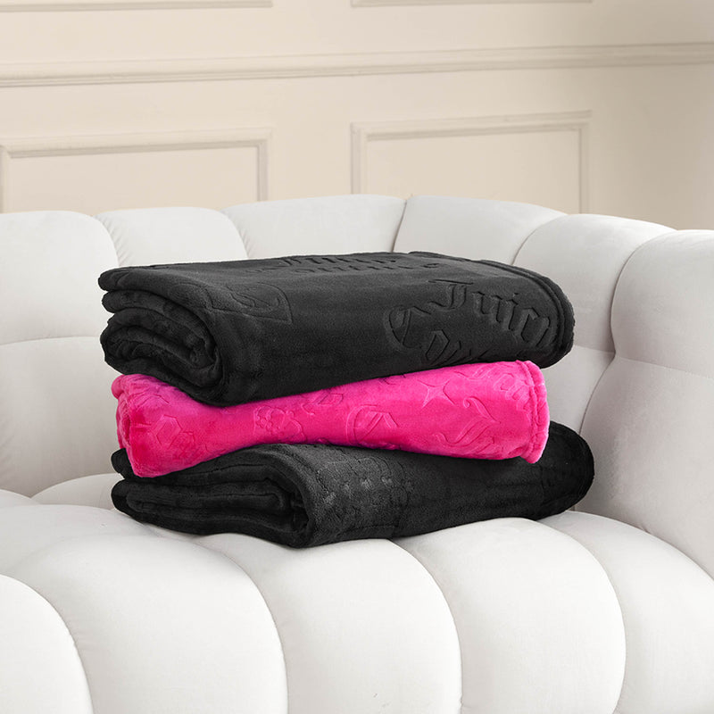 Three folded blankets—two black and one bright pink Embossed Plush Throw Blanket—are stacked on a white, tufted sofa in front of a beige wall with decorative molding.