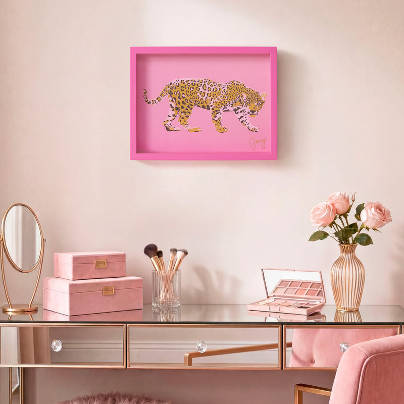 A vanity table adorned with pink makeup items, a vase of pink roses, and a mirror sits below the elegant Shadow Box Wall Art, 12″ x 16″. The framed artwork features a chic pink leopard design by Juicy Couture, adding a fashionable touch that blends seamlessly into the feminine décor.