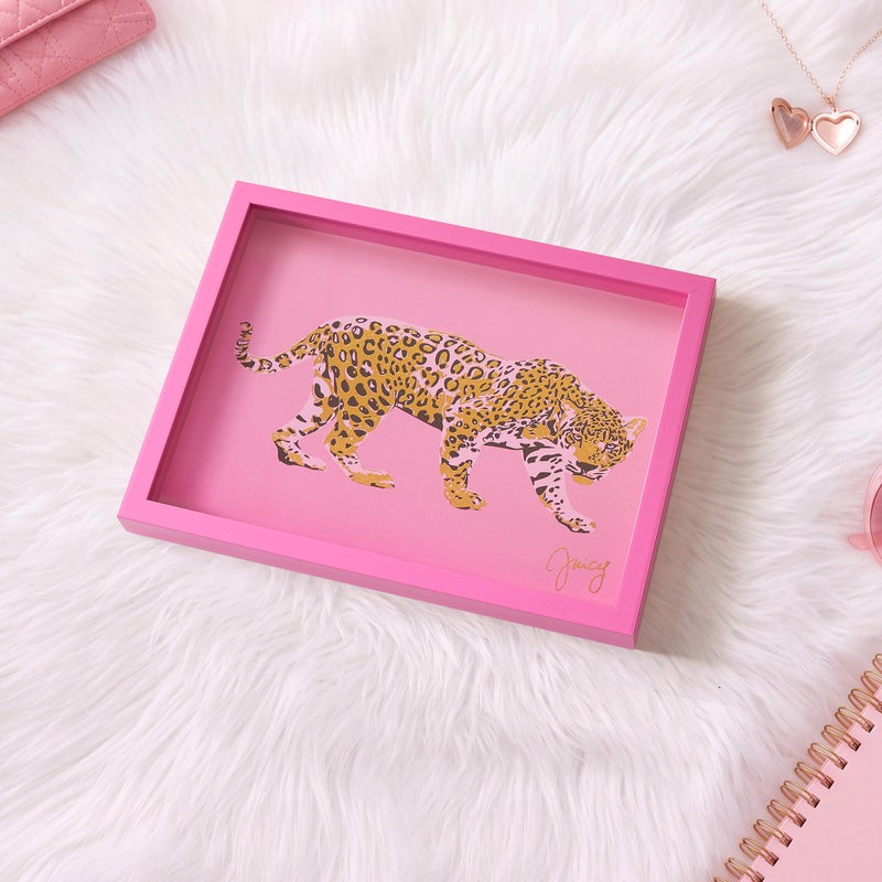 A pink-framed Shadow Box Wall Art (12″ x 16″) featuring an illustration of a leopard on a pink background sits on a white fur surface, surrounded by pink accessories and a spiral notebook. This chic piece channels fashion-inspired artwork, adding a playful yet stylish accent to any space.