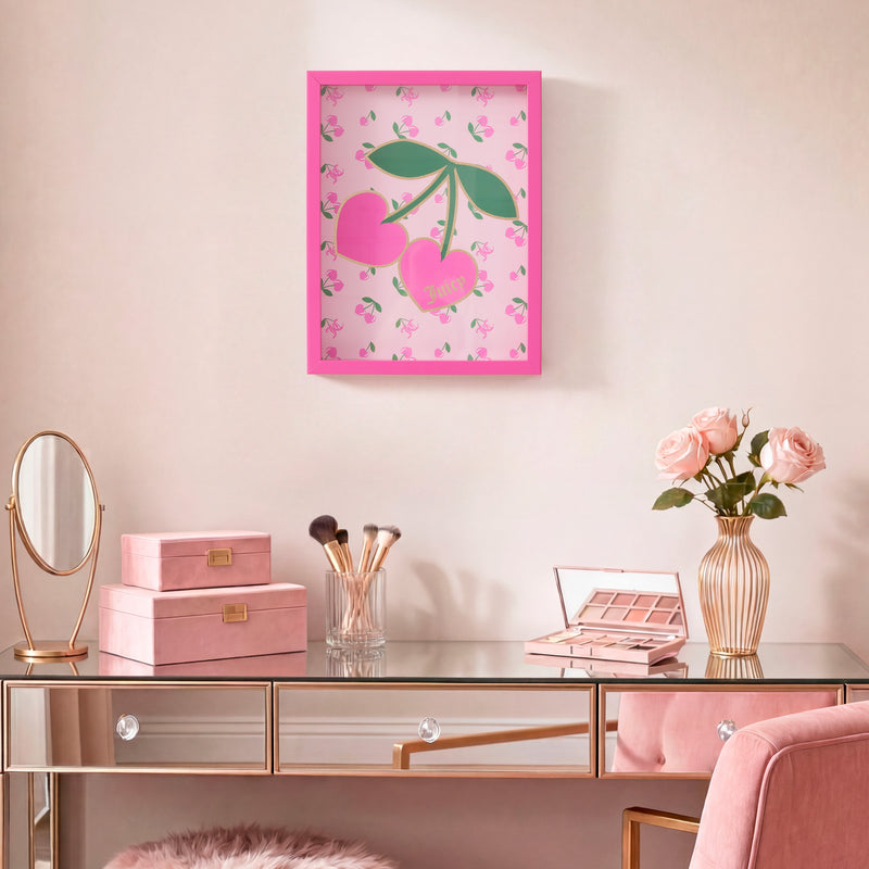 A chic vanity with a glass surface holds makeup brushes, a mirror, and pink boxes. Above, a framed Shadow Box Wall Art (12″ x 16″) hangs on the pale pink wall, adding a touch of fashion-inspired decor to the scene. A vase of pink roses brings a fresh element to the stylish, feminine space.