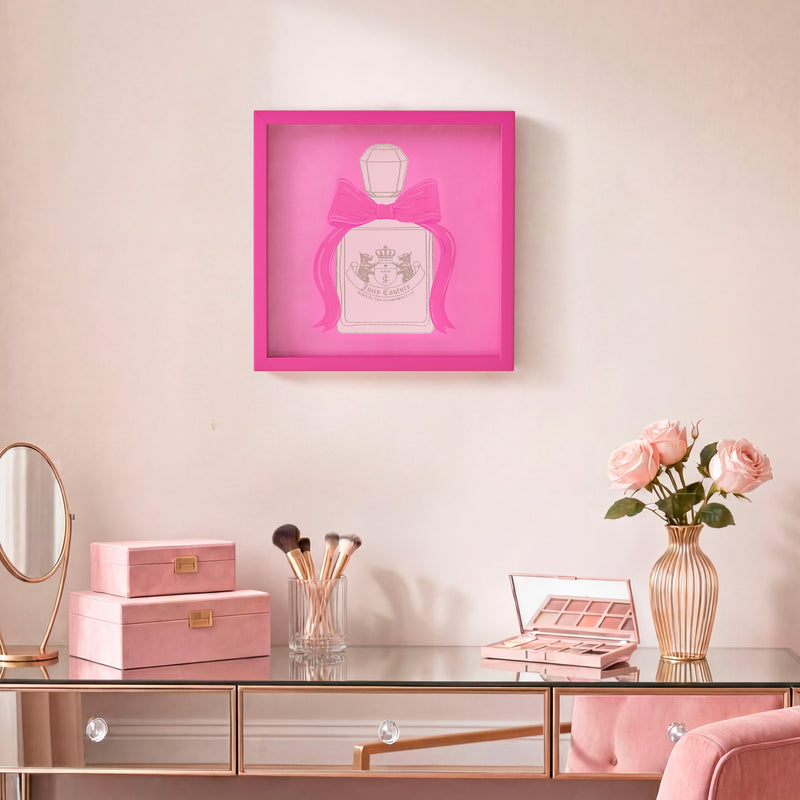 A vanity with makeup brushes, pink boxes, a palette, and a vase of roses. Above it hangs the Shadow Box Wall Art (14″ x 14″), featuring pink perfume bottle artwork with a bow—bringing a touch of Juicy Couture-inspired style to the space.