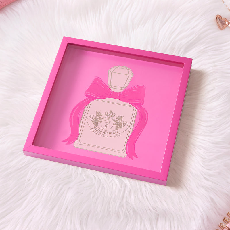 A 14″ x 14″ Shadow Box Wall Art featuring an illustration of a perfume bottle with a large pink bow, set against a white furry background. Accented with rose gold heart paper clips, this fashion-inspired piece makes for an adorable Juicy Couture-themed decor addition.