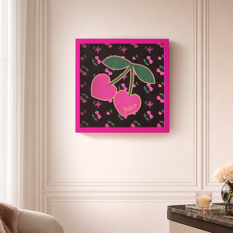 A 14″ x 14″ Shadow Box Wall Art featuring two pink cherries—one labeled “Juicy”—is displayed on a beige wall above a table with a candle and a book. This fashion-inspired piece showcases a black background patterned with small pink cherries and logos, adding a stylish touch to any space.