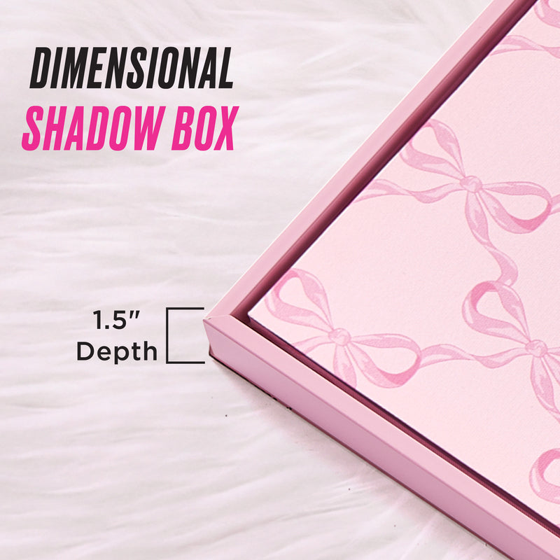 A Shadow Box Wall Art, 16″ x 20″, with a 1.5-inch depth and pink bow pattern is displayed on a white fur surface. The bold text “Dimensional Shadow Box” is featured on the piece, offering a chic, fashion-inspired touch that beautifully elevates your space—ideal for fans of Juicy Couture wall art aesthetics.