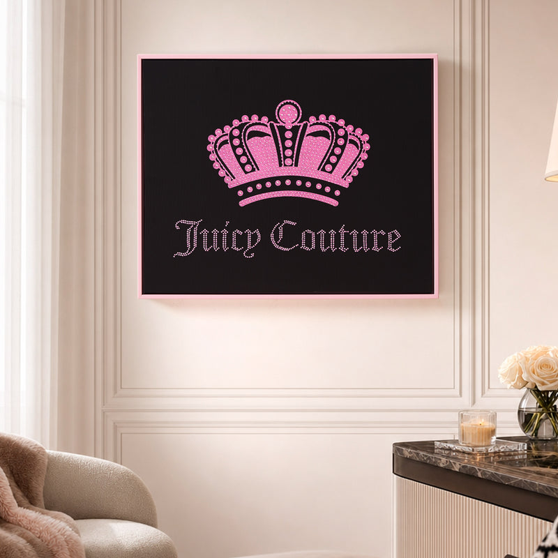 A Shadow Box Wall Art, 16″ x 20″, featuring a pink crown above the words “Juicy Couture” in pink lettering, hangs on a cream-colored wall above a marble table adorned with a vase of white roses. This fashion-inspired piece brings a chic statement to the space.