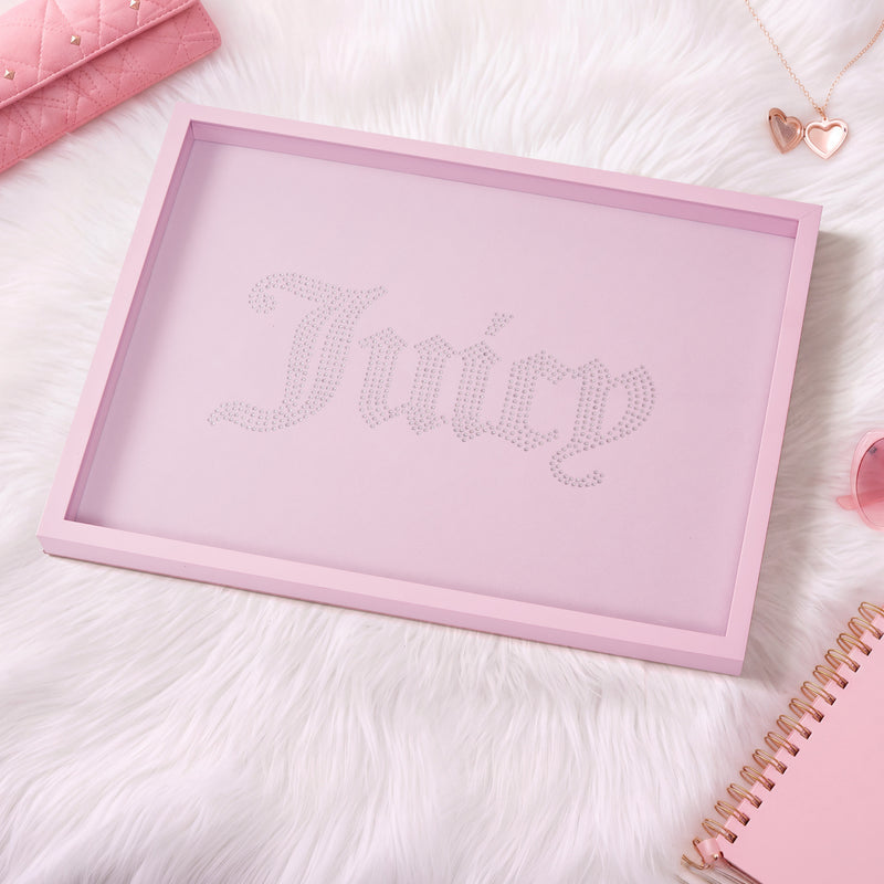 A pink-framed Shadow Box Wall Art (18″ x 24″) featuring the word “Juicy” in rhinestones sits atop a white fur surface, surrounded by pink accessories, heart-shaped jewelry, and a notebook—making it the perfect fashion-inspired decor for any chic space.