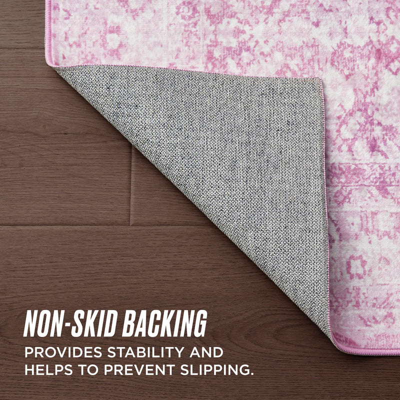 A Finely Washable Rug, 3' x 5', in a pink pattern rests on a wooden floor with one corner flipped to show its textured gray non-skid backing. Text: NON-SKID BACKING provides stability and helps prevent slipping.