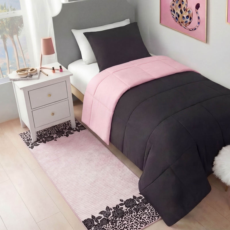 A neatly made twin bed with a dark gray comforter and pink accents sits beside a white nightstand topped with a pink lamp and beauty items. Next to the bed, beneath leopard art, lies the Finely Washable Rug, 2' x 6', featuring ultra-soft pink and black florals.