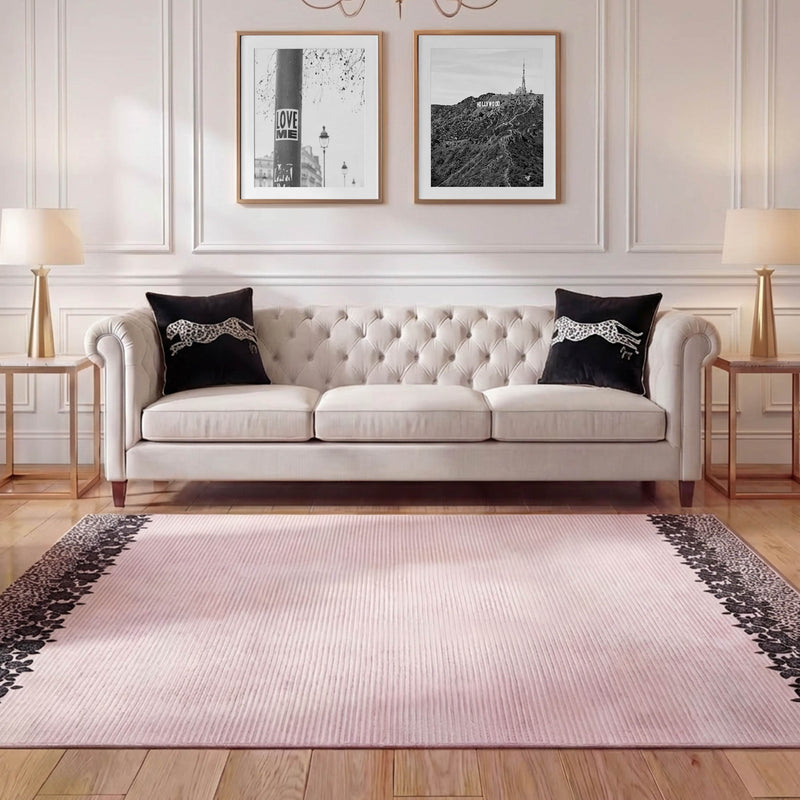 A beige tufted sofa with black and white pillows sits against a white wall with framed photos. Side tables with lamps flank the sofa, while a Finely Washable Rug, 7'10 x 10', featuring dark borders covers the wooden floor.