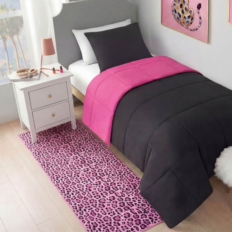 A neatly made bed with black and bright pink bedding stands by a white nightstand with cosmetics and a lamp. A Finely Washable Rug, 2' x 6', in pink leopard print covers the floor, matching the pink leopard artwork above the bed.