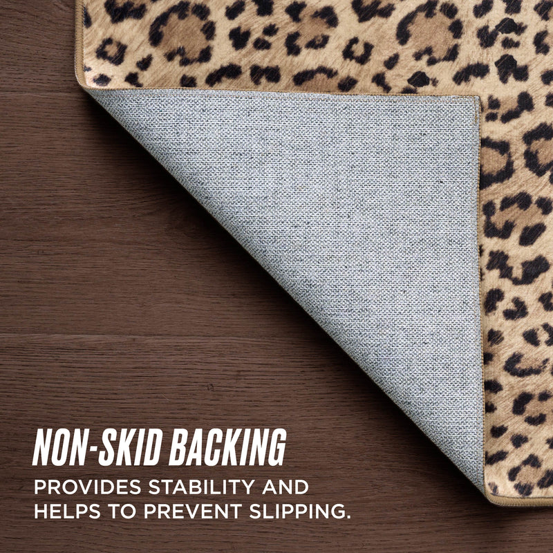 A corner of the Finely Washable Rug, 5' x 7', with leopard print is lifted to show its non-skid backing on a dark wood floor. Text overlay: NON-SKID BACKING provides stability and helps prevent slipping.