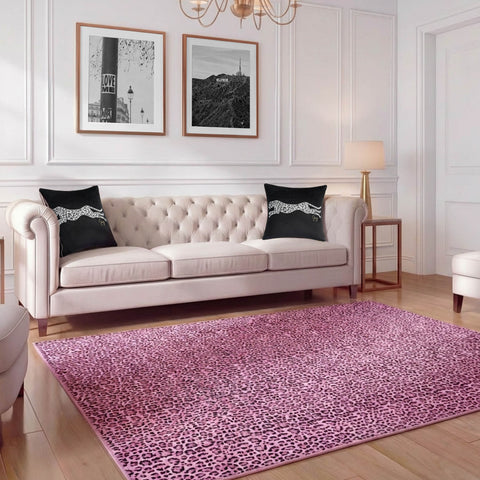 A modern living room with a tufted beige sofa, black cheetah throw pillows, a pink leopard print Finely Washable Rug (7'10 x 10') with non-skid backing, two black-and-white photos above the sofa, white walls, and gold accents.