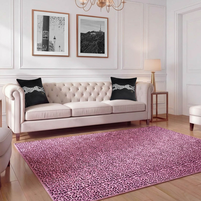A modern living room with a tufted beige sofa, black cheetah throw pillows, a pink leopard print Finely Washable Rug (7'10 x 10') with non-skid backing, two black-and-white photos above the sofa, white walls, and gold accents.