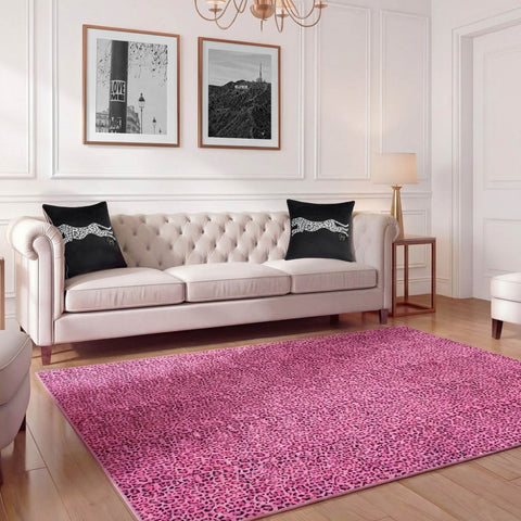 A stylish living room features a white tufted sofa, black cheetah-print pillows, two black-and-white framed photos, and a Finely Washable Rug, 7'10 x 10', in pink leopard print under a gold floor lamp on warm wooden flooring.