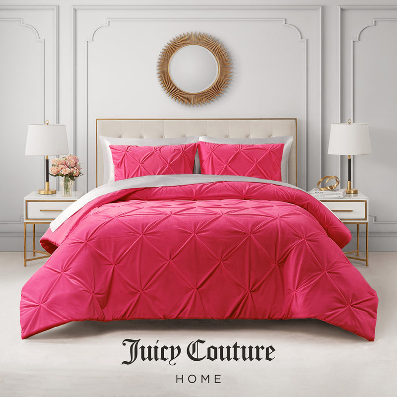 A modern bedroom showcases the Kiss Pleat Micromink Comforter Set in bright pink, paired with white nightstands, gold lamps, a vase of roses, and a sunburst mirror. "Ultra-luxurious bedding by Juicy Couture Home" appears at the bottom.