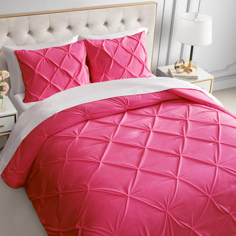 A neatly made bed with a tufted beige headboard, the Kiss Pleat Micromink Comforter Set in bright pink with matching pillows, white sheets, a white bedside lamp, and a gold and white nightstand decorated with accents.